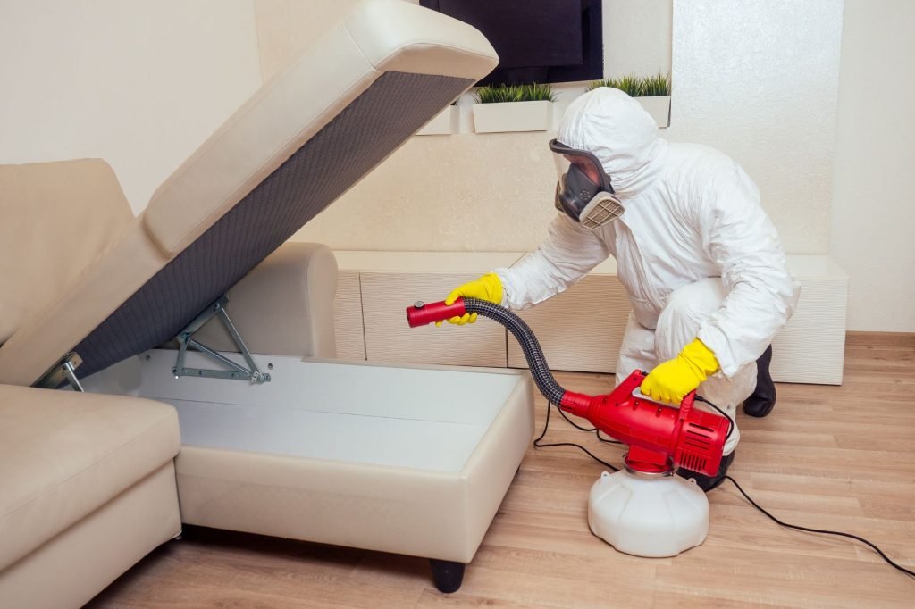 A man in protective gear and a white suit uses a vacuum to clean a couch for bed bug control in Honolulu, HI.